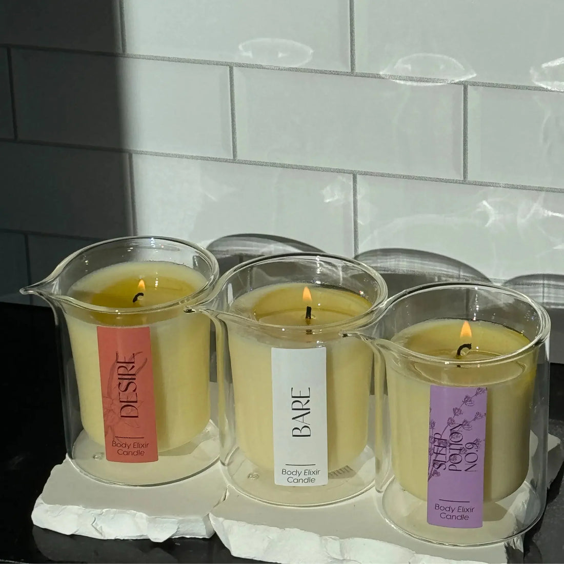 Three lit Body Elixir Candles in clear glass holders labeled Desire, Bare, and Super Potion No.9 on white stone slabs