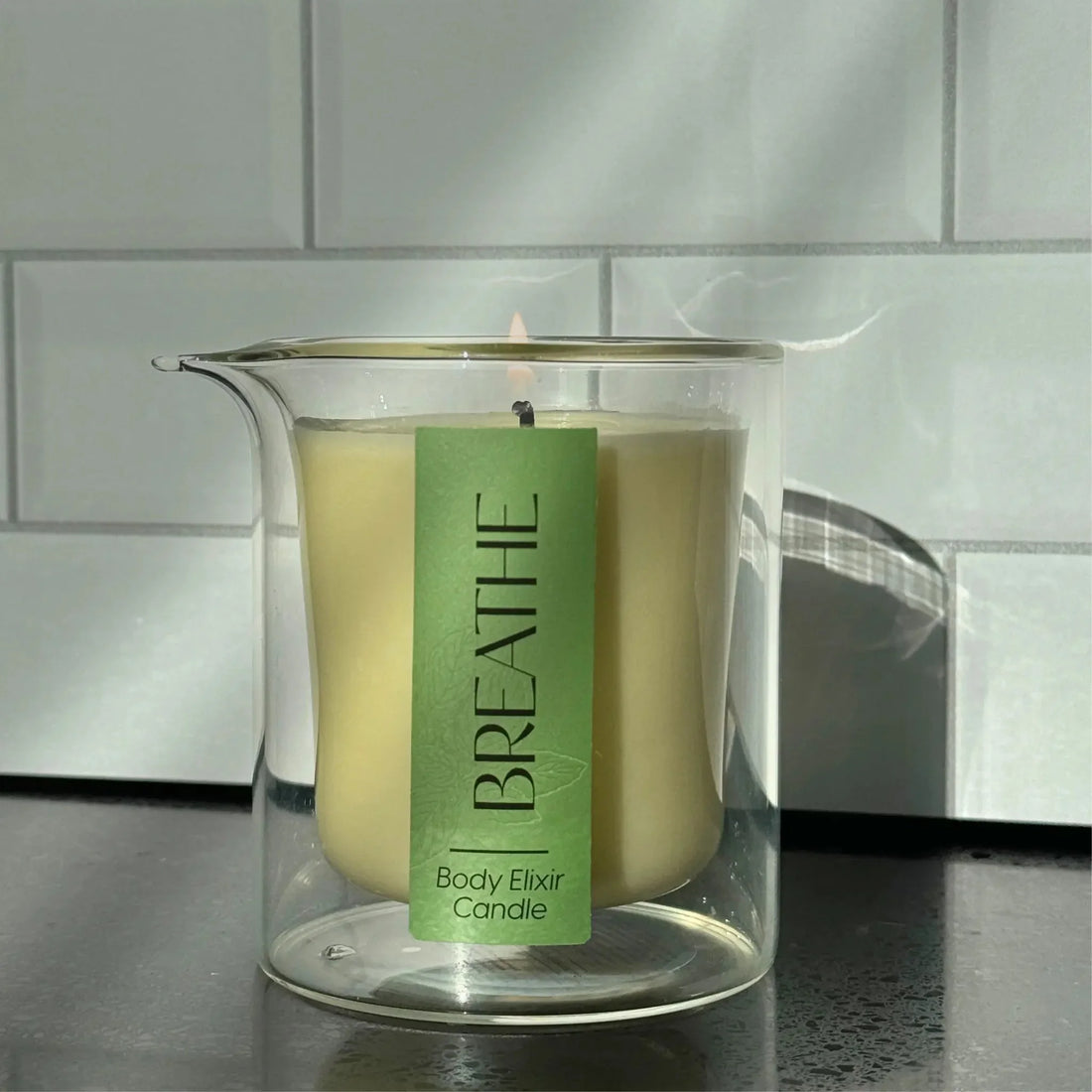 Lit BREATHE Body Elixir Candle in clear glass container with spout on black countertop against white tiled wall