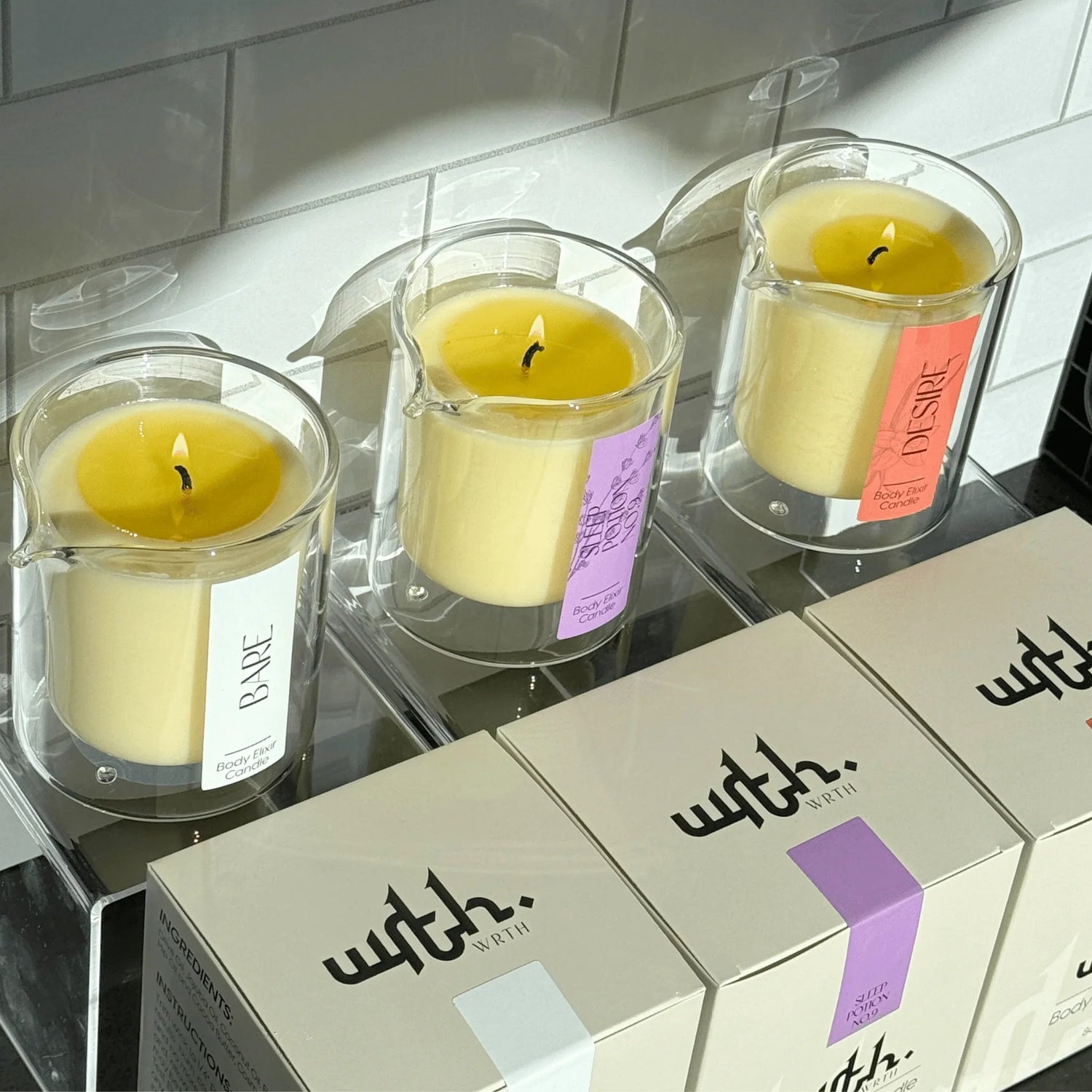 Three lit WRTH body elixir massage oil candles in glass holders labeled Bare, Sleep Potion No.9, Desire with matching branded boxes