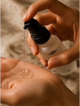 Hand holding a white bottle with a black cap, dripping clear liquid onto another hand.