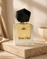 A frosted glass perfume bottle labeled 'with. Rose Oud' with pale yellow liquid, a black faceted cap, and gold lettering, displayed on a stone slab in a warm neutral setting.