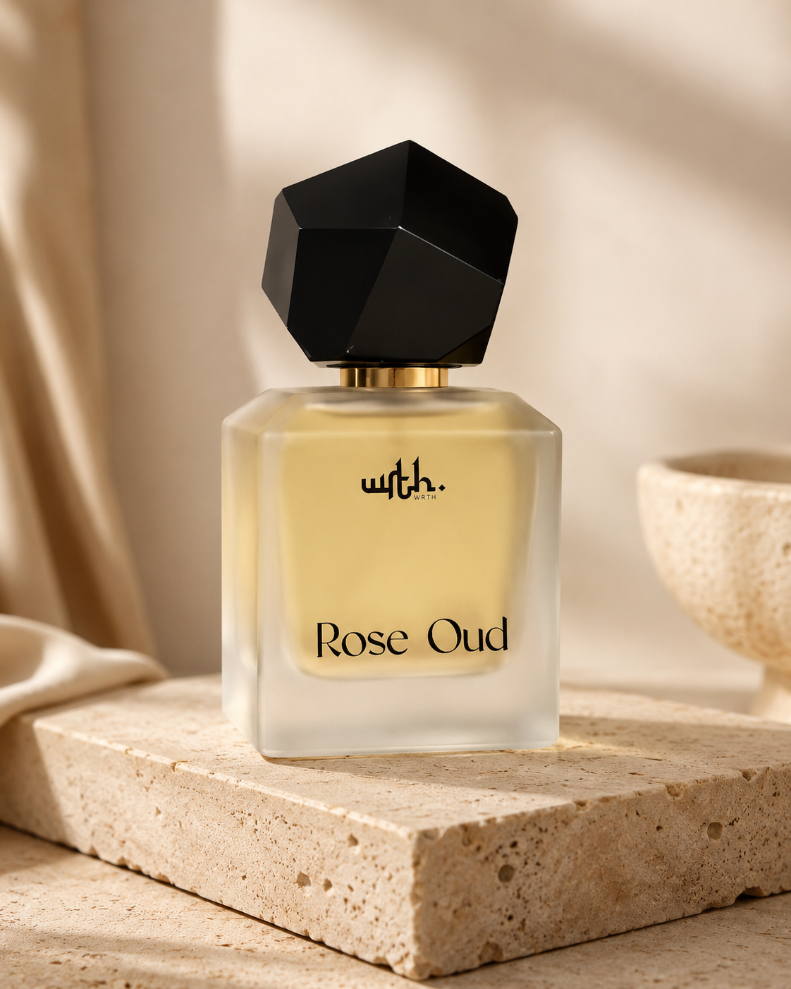 A frosted glass perfume bottle labeled 'with. Rose Oud' with pale yellow liquid, a black faceted cap, and gold lettering, displayed on a stone slab in a warm neutral setting.