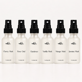 Six small white spray bottles with black pumps arranged in a row on a light gray background. The bottles are labeled with different fragrance names: Cherry Musk, Rose Oud, Opulence, Vanilla Musk, Mango Musk, and Jasmine Musk.