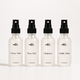 Four white spray bottles with black caps on a white background, labeled with different scents.