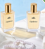 Two clear rectangular perfume bottles with gold caps and pale yellow liquid, labeled 'wrth Opulence,' arranged on a white surface with a pearl strand in the foreground and a soft blue background.