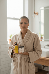 Person in beige bathrobe holding WRTH.Studio vanilla-scented body oil spray and sugar scrub in bright, minimalist bathroom