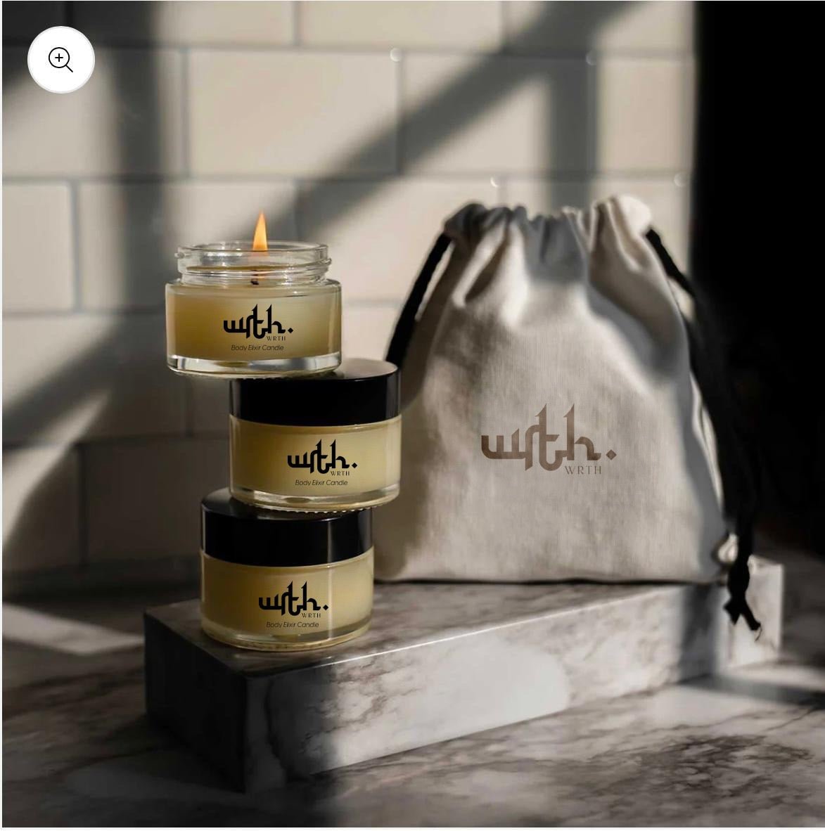 Stack of three WRTH body elixir candles, one lit, beside a branded drawstring pouch on a marble surface