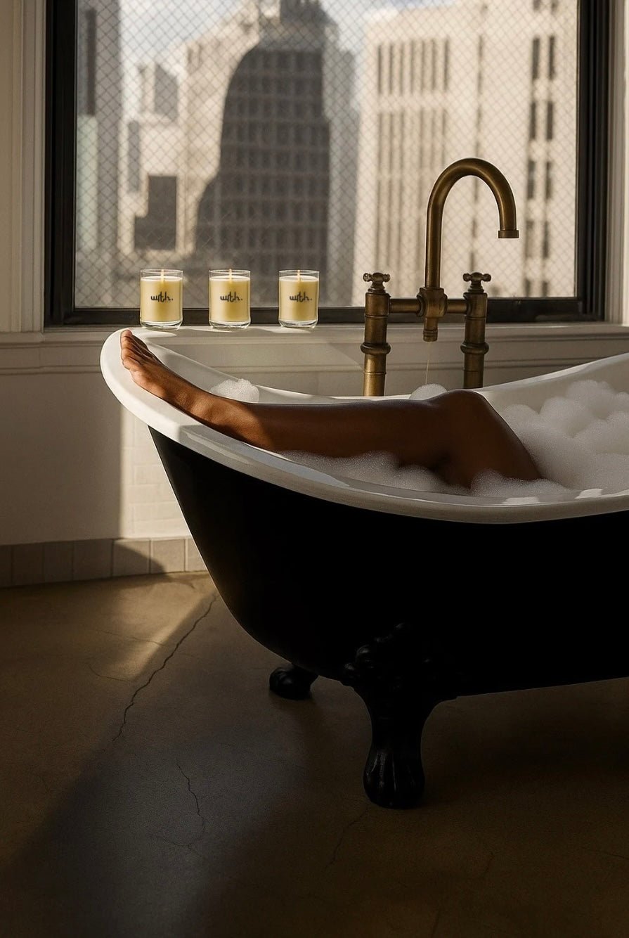 Person relaxing in black clawfoot bathtub with bubbles and three lit WRTH candles by large window
